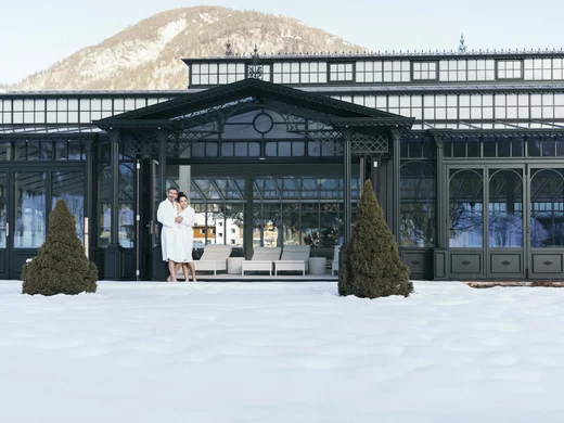 Wiesenhof am Achensee | Das Hotel in Pertisau Paar in Bademänteln vor einem Winterhotel mit verschneitem Garten und Bergen
