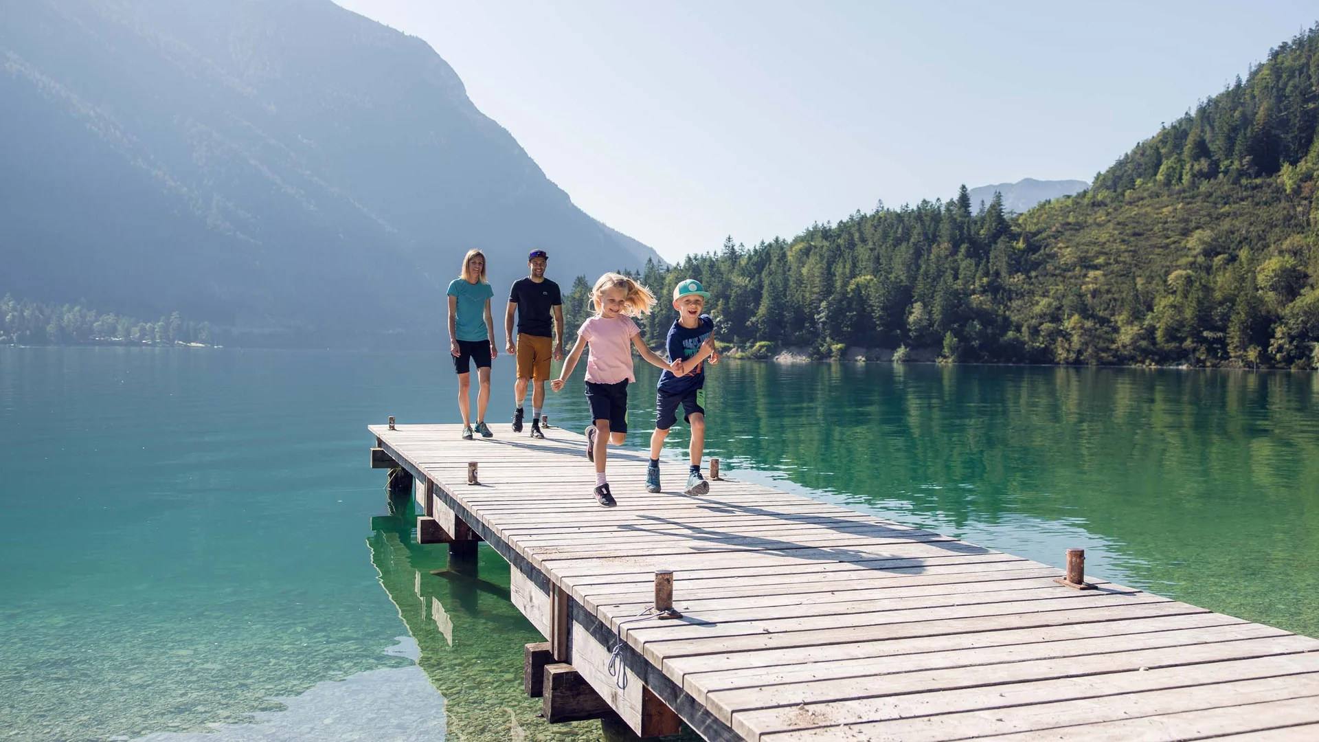 Discover Lake Achensee Family with two children running on wooden pier over clear mountain lake surrounded by mountains