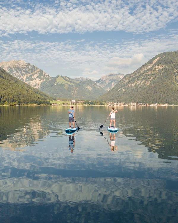 Wiesenhof am Achensee | Das Hotel in Pertisau Zwei Personen auf Stand-up-Paddle-Boards auf einem stillen Bergsee bei bewölktem Himmel