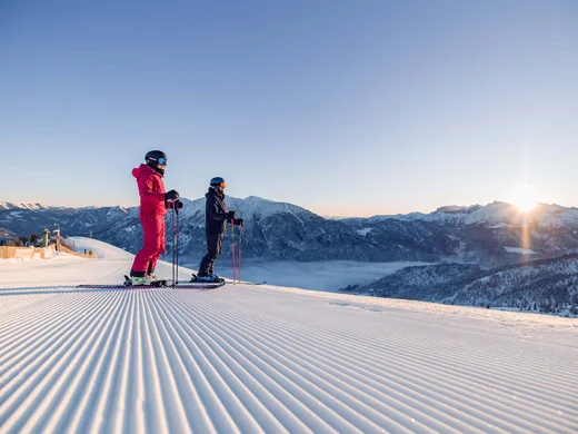 Discover Lake Achensee Two skiers standing on a groomed slope overlooking snowy mountains at sunrise